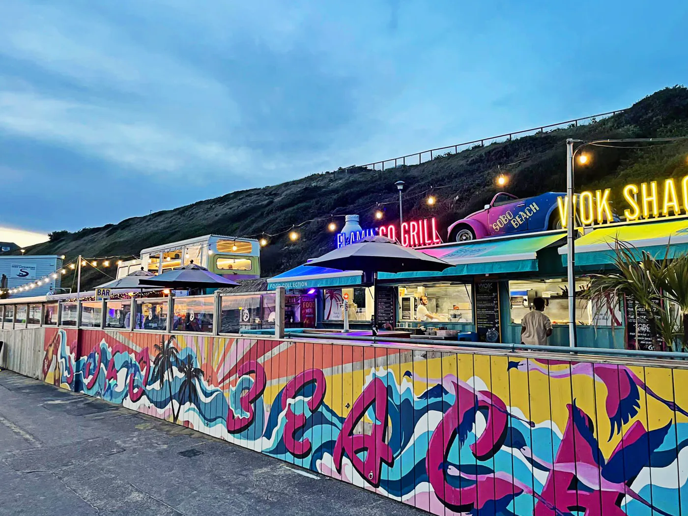 SOBO Beach at dusk, with a painted beach mural along the fence, festoon lights and neon signs for the Flamingo Grill and Wok Shack
