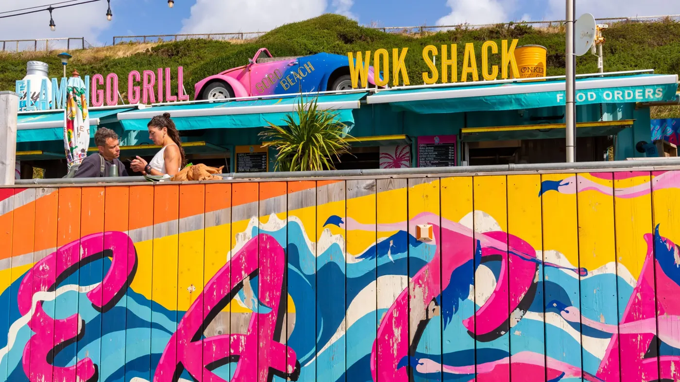 Two people talking at a table behind a brightly painted fence at SOBO Beach, under the Flamingo Grill and Wok Shack signs